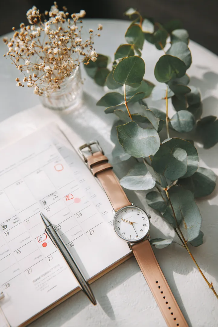 Flat lay of a monthly wall planner with circled due dates beside a wristwatch and eucalyptus sprigs